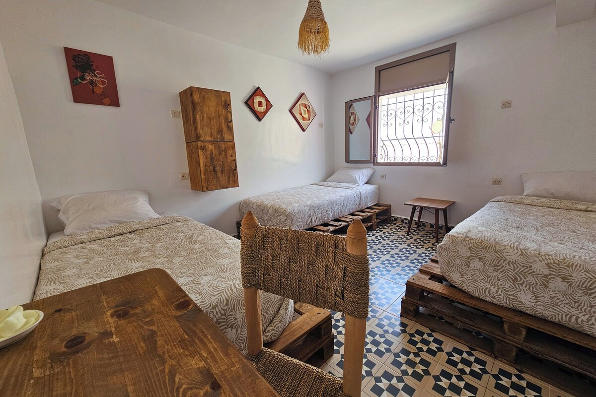 Una habitación con dos camas, una mesa de madera, una silla de fibras naturales y una lámpara colgante.
