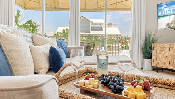 Ein Zimmer mit Blick auf einen Strand, eine Couch, ein Tablett mit Käse und Obst sowie eine Weinflasche.