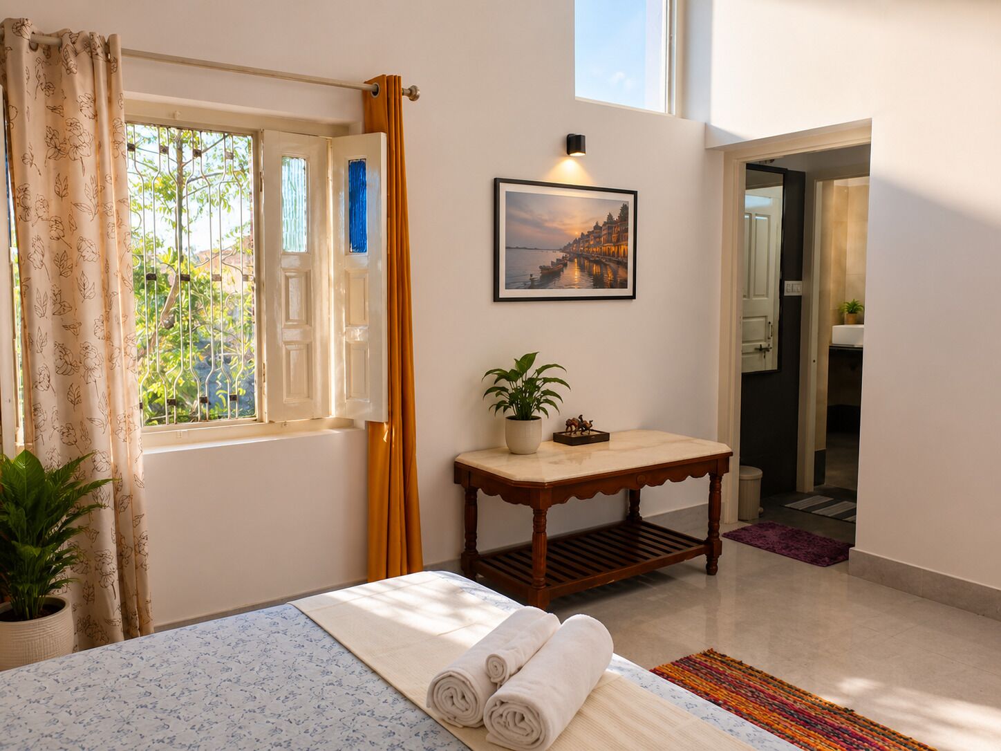 A hotel room with a bed, a table with a plant, a window with curtains, and a painting on the wall.
