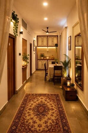 A hallway with a patterned rug, wooden doors, a ceiling fan, and a dining area with chairs and a table. - Hotel Atithya (Udaipur)