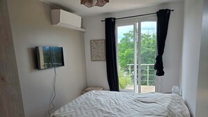A bedroom with a bed, a TV mounted on the wall, a window with curtains, and a balcony with a railing.