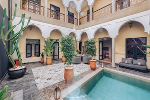 Un área de piscina con suelo de baldosas, plantas en macetas y una zona de estar con cojines.
