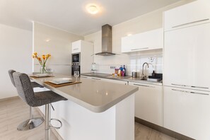 A modern kitchen with white cabinetry, a central island, bar stools, a built-in oven, and a stainless steel range hood. - Casa Foti Puerto del Carmen - Media estancia (Puerto del Carmen)