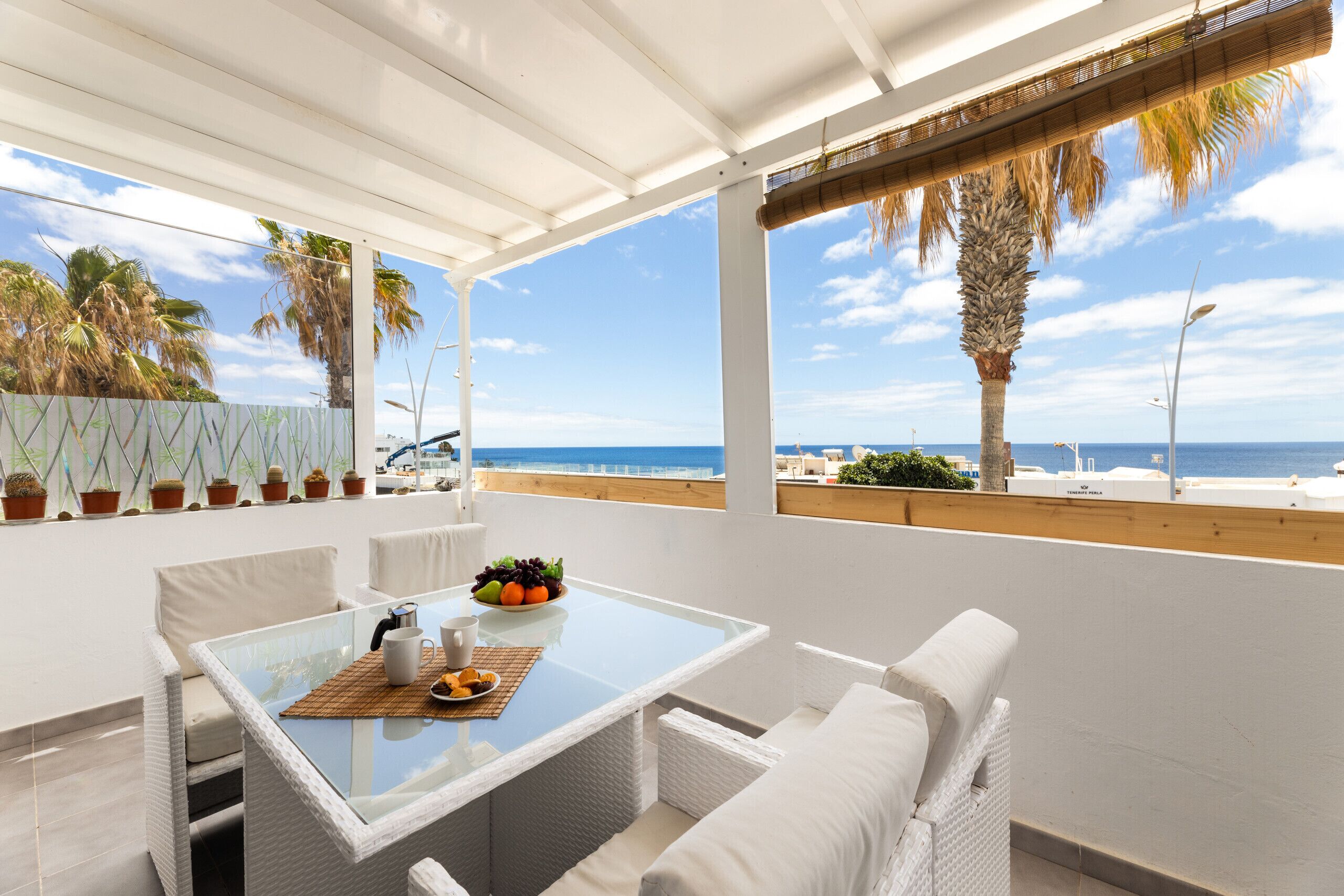 Ein Balkon mit Glastisch, weißen Korbmöbeln und Blick auf das Meer und Palmen.