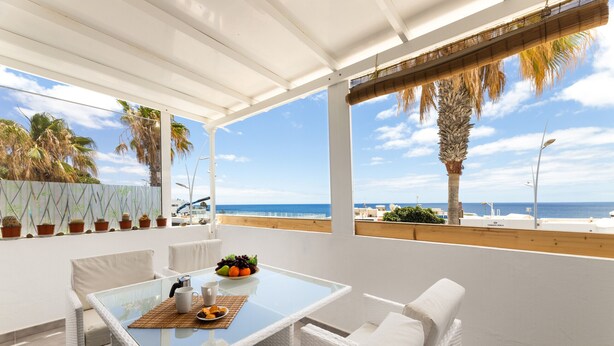 En balkon med et glasbord, hvide fletmøbler og udsigt til havet og palmer.