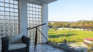 A balcony with a telescope, a wicker sofa, and a view of a green field and distant buildings.