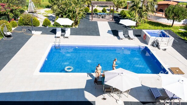 A rectangular pool with two lounge chairs and a sun umbrella, surrounded by a tiled deck and a pergola.