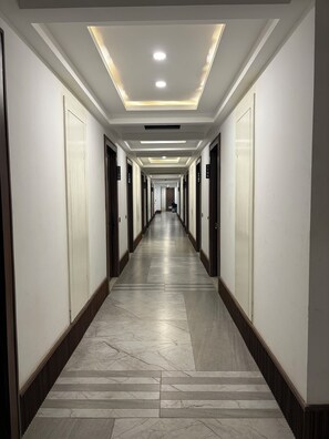 Un long couloir étroit, avec des portes des deux côtés, chacune portant un numéro. Le sol est recouvert de carreaux de marbre formant un motif, et le plafond est doté d’un éclairage encastré.