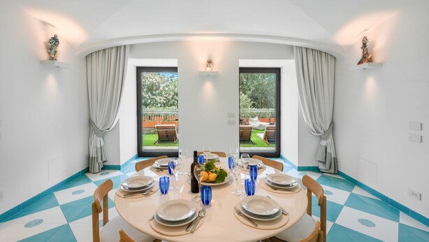 Ein Essbereich mit einem runden Tisch für vier Personen, großen Fenstern mit Vorhängen und Blick auf einen Garten.