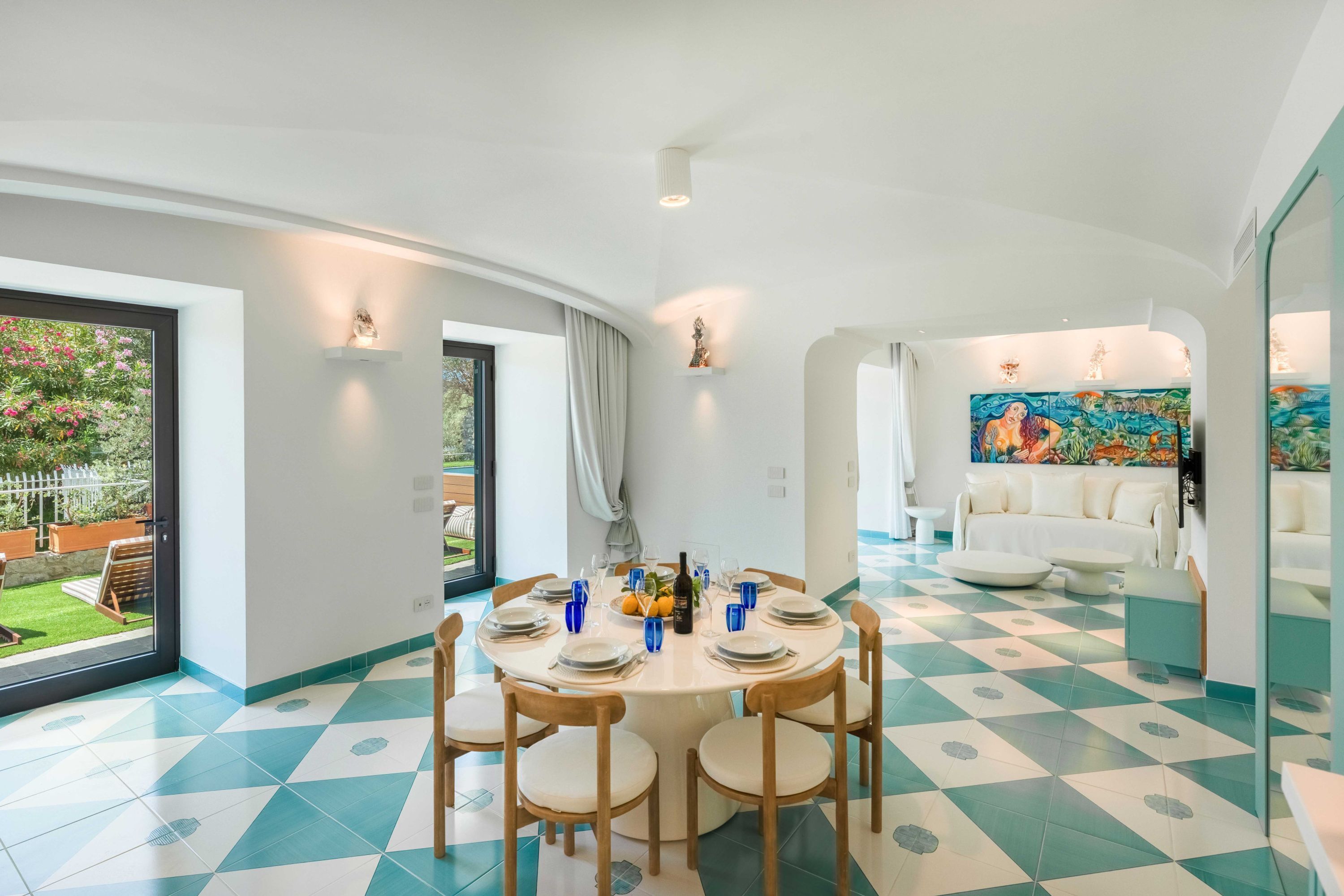 Un coin repas avec une table ronde dressée pour quatre personnes, un canapé et une fresque colorée en arrière-plan.