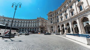 Ein gepflasterter Platz mit einem historischen Gebäude, das Rundbogenfenster und Säulen aufweist, einem Bus und parkenden Autos.