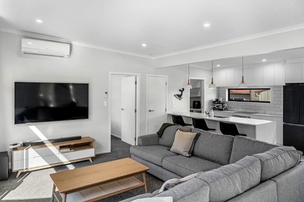 A modern living room with a grey sofa, a wooden coffee table, a flat-screen TV, and a kitchen area with white cabinets and a black refrigerator.