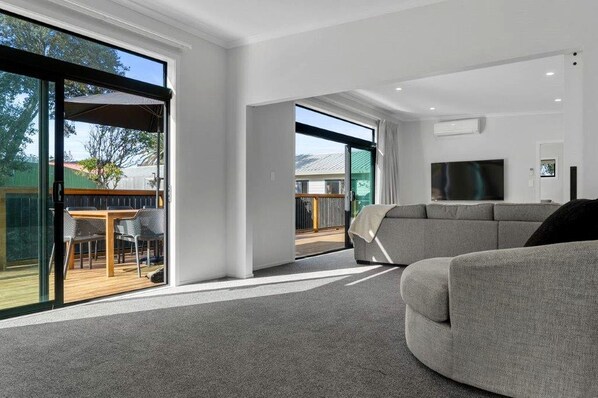 A modern living room with a grey sofa, a flat-screen TV, and a glass door leading to an outdoor area with a table and chairs.