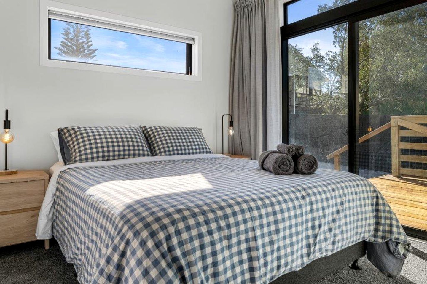 Une chambre avec un grand lit, une literie à carreaux, une table de chevet, une lampe et une vue sur une terrasse en bois et des arbres.