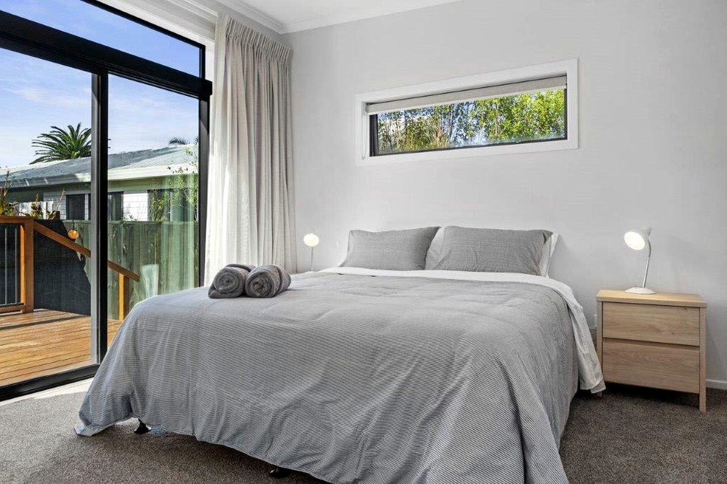 A bedroom with a bed, two bedside tables, a window, and a view of a deck.