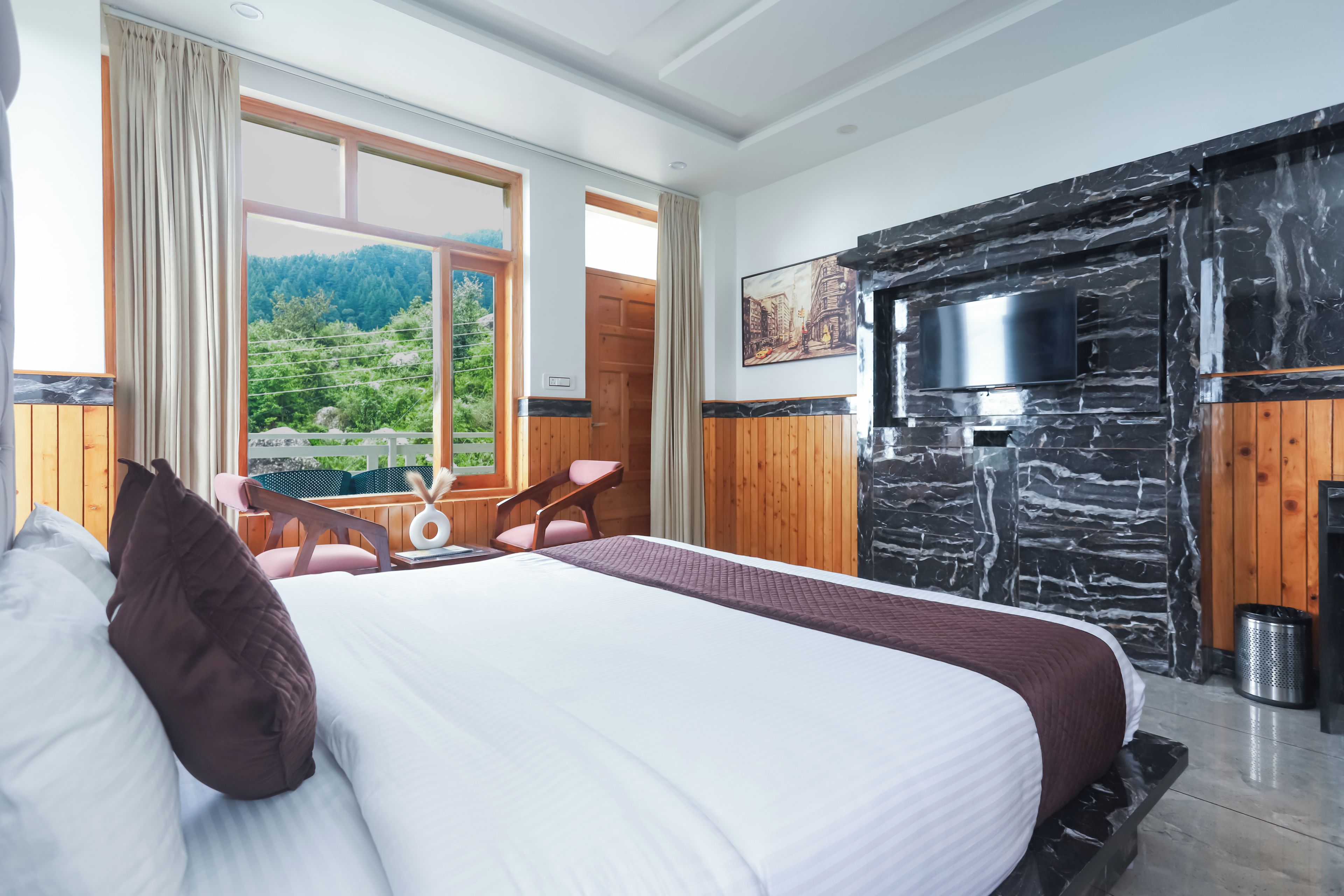A bedroom with a bed, a TV mounted on a stone wall, a window with a view of greenery, and a wooden door.