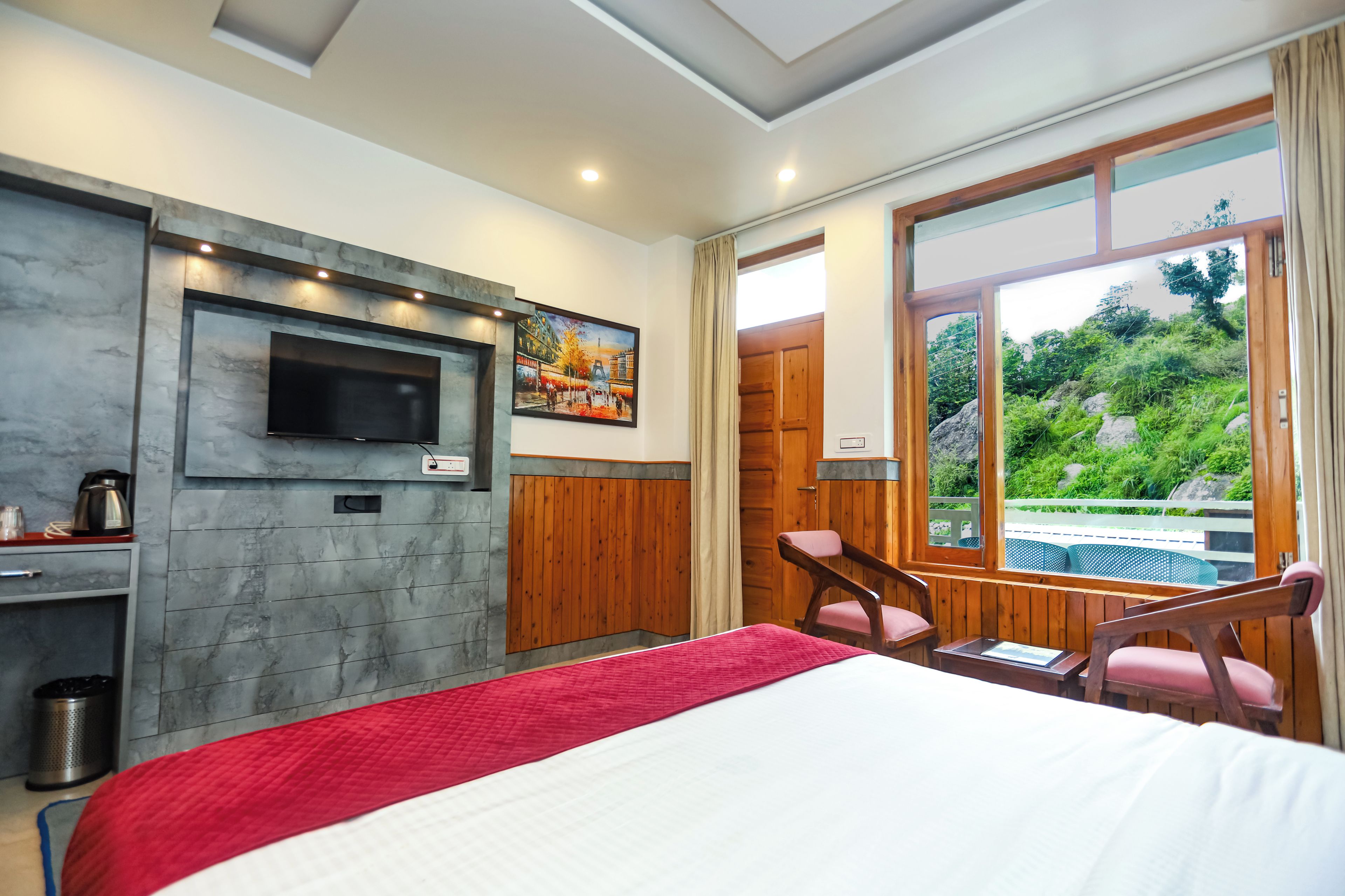 Habitación de hotel con cama, televisor montado en la pared, un cuadro, una puerta de madera y vistas al verdor exterior.