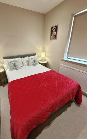 A bedroom with a bed, a red blanket, a nightstand with a lamp, and a window with blinds. - Modern Two-Bed Apartment in Central Bath (Bath)