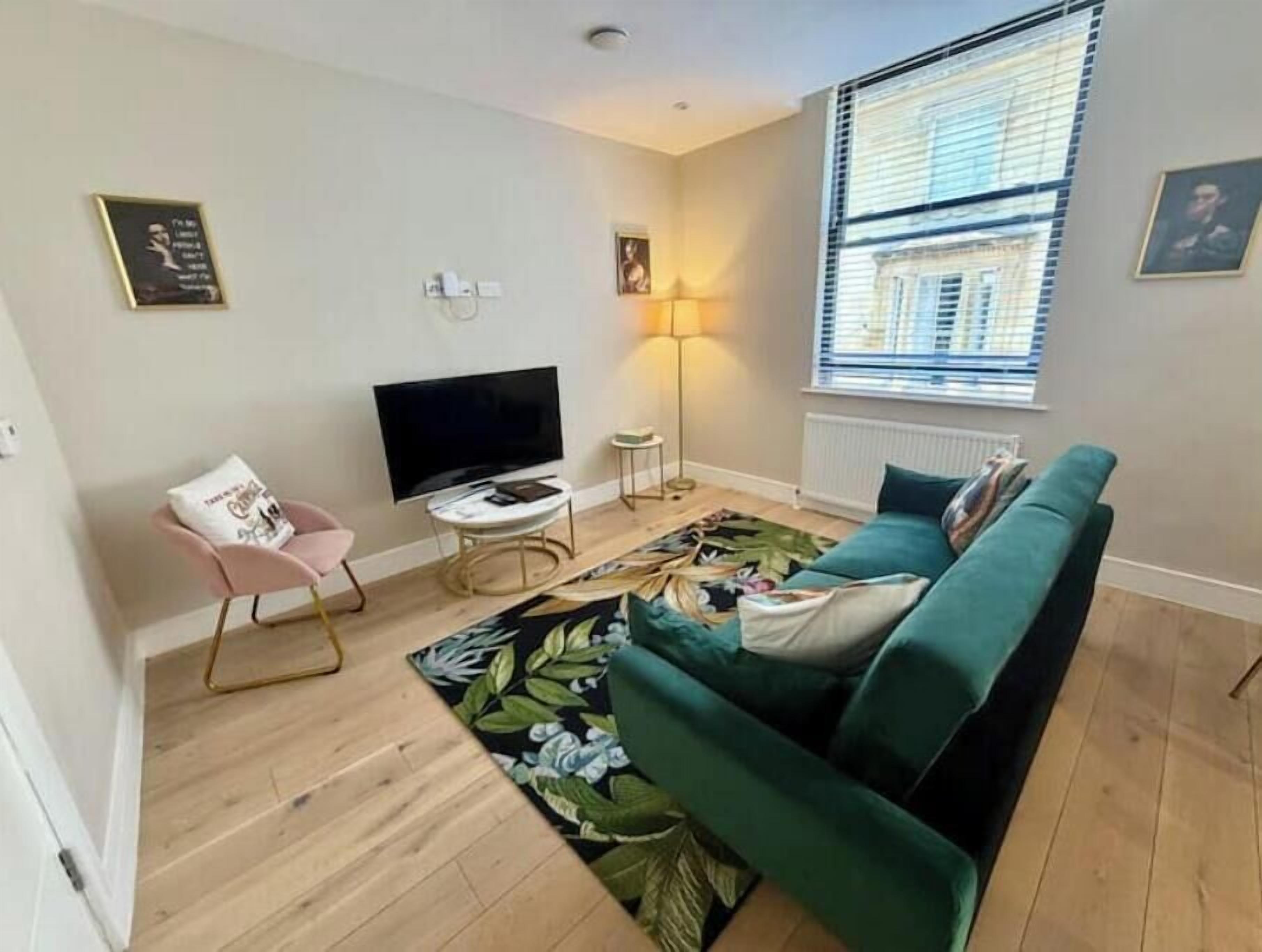 A living room with a green sofa, a flat-screen TV, a pink chair, a floor lamp, and a window with blinds.