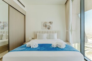 A hotel room with a bed, a blue blanket, rolled towels, a nightstand, a lamp, and artwork on the wall. - PVH - Binghatti Royale 1BR Private Pool (Dubai)