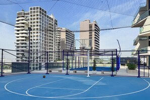An outdoor basketball court with a blue surface, surrounded by a chain-link fence, and high-rise buildings in the background. - PVH - Binghatti Royale 1BR Private Pool (Dubai)