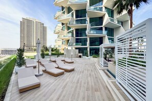 A rooftop lounge area with sun loungers, a palm tree, and a modern building in the background. - PVH - Binghatti Royale 1BR Private Pool (Dubai)