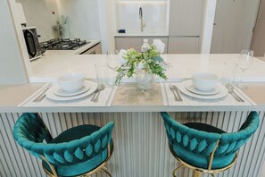 A modern kitchen with a central island, two teal upholstered chairs, and a decorative vase with flowers. - PVH - Binghatti Royale 1BR Private Pool (Dubai)