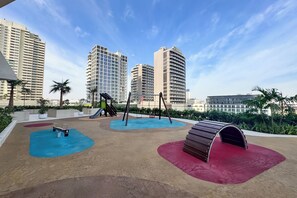 A playground with swings and slides, surrounded by palm trees and buildings. - PVH - Binghatti Royale 1BR Private Pool (Dubai)