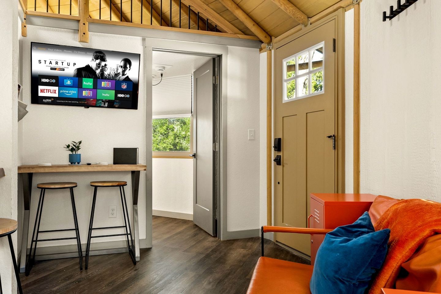 Ett kompakt bostadsutrymme med en TV som är monterad på väggen och visar strömnings­tjänster, ett litet köksområde med pallar och en dörr som leder ut till ett utomhusområde.
