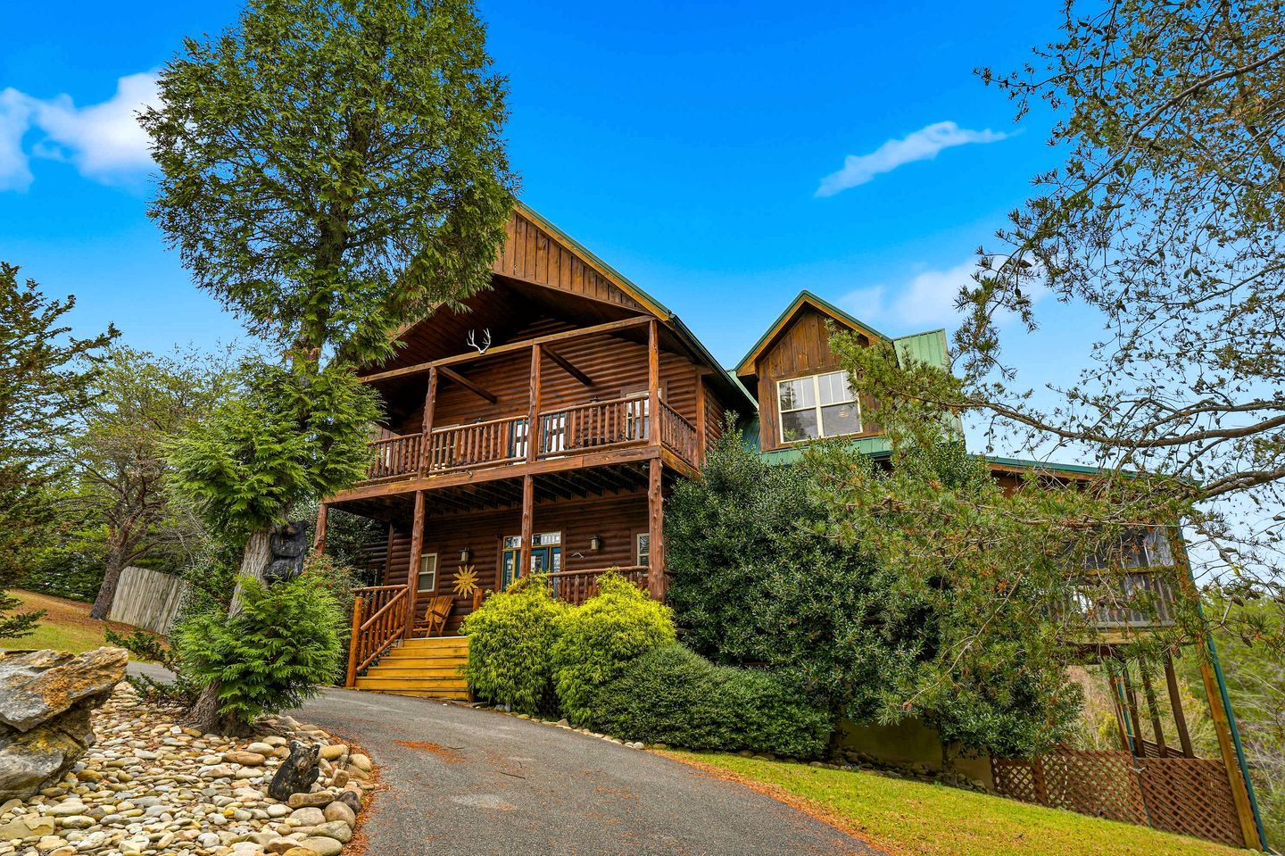 A two-story log cabin with a balcony, surrounded by dense greenery and a stone pathway.