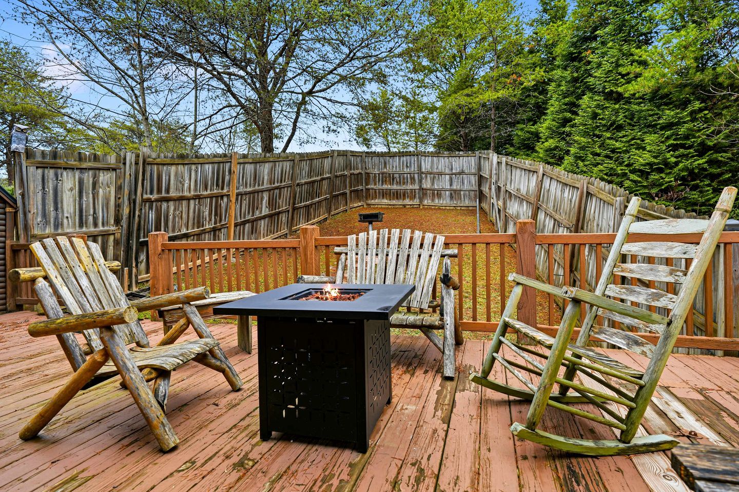 A wooden deck with a fire pit table, two Adirondack chairs, and a wooden fence.