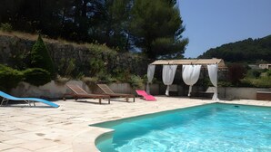 En swimmingpool med stengærde, liggestole og en pergola med hvide gardiner.