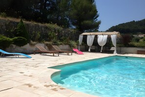 A swimming pool with a stone border, lounge chairs, and a pergola with white curtains. - Holiday Home 'Bergerie Des Estelles' with Mountain View, Wi-Fi and Air Conditioning (Auriol)
