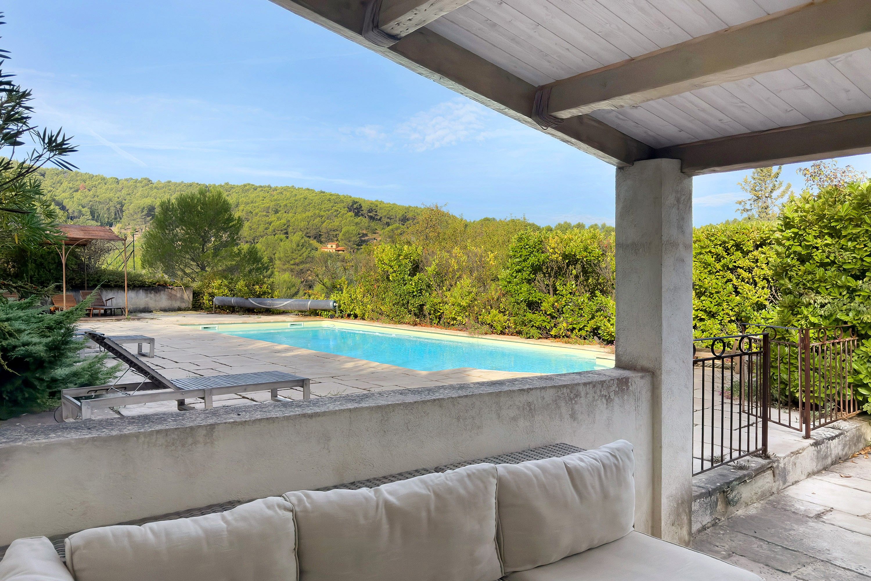 A covered outdoor area with a swimming pool, lounge chairs, and a view of a hillside.