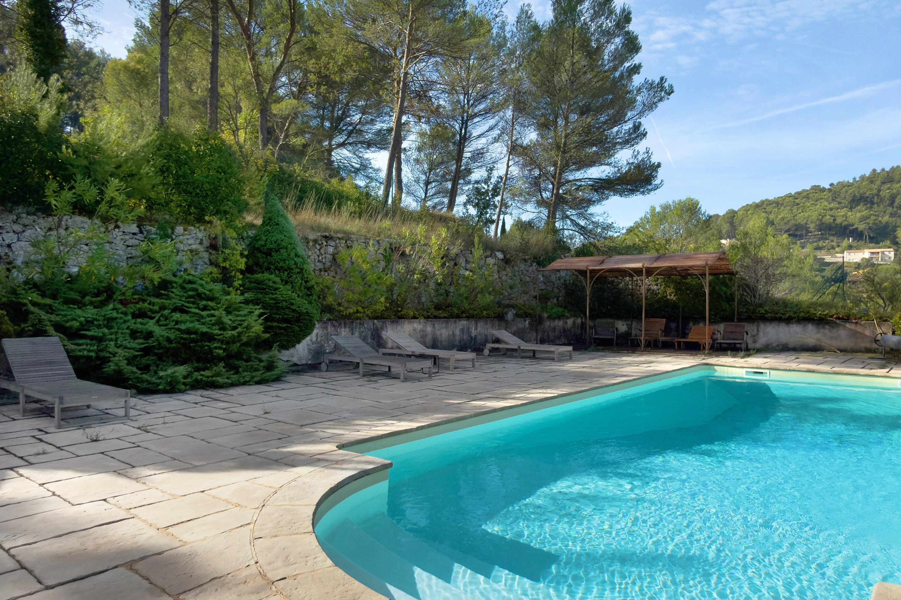 Piscina com deck de pedra, rodeada por árvores e um muro de pedra.