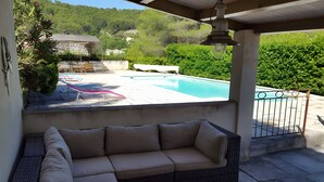 Et overdækket udendørs opholdsområde med en pool, en stenvæg og grønt i baggrunden.