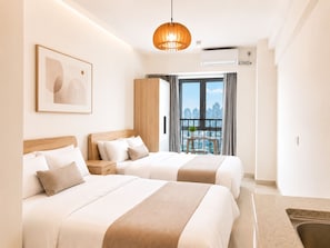 A hotel room with two beds, a large window with a city view, a hanging light fixture, and an air conditioning unit.