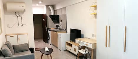 A compact hotel room with a bed, a small desk, a chair, a sofa, a kitchenette, and a wardrobe.