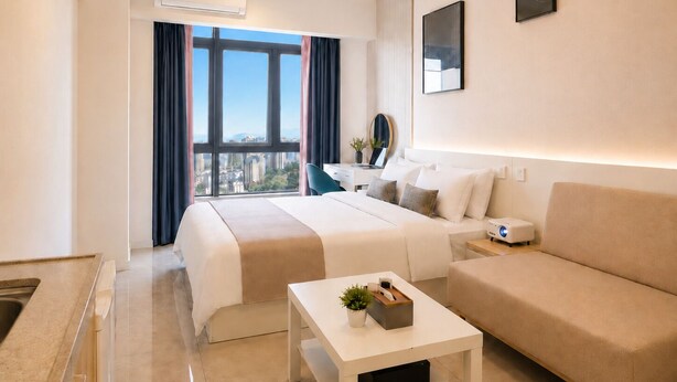 A modern hotel room with a bed, a sofa, a small table, and a desk. There is a window with curtains, an air conditioning unit, and a ceiling light fixture. -  Nestory Residence - Foshan Lecong Furniture Market  (Foshan)