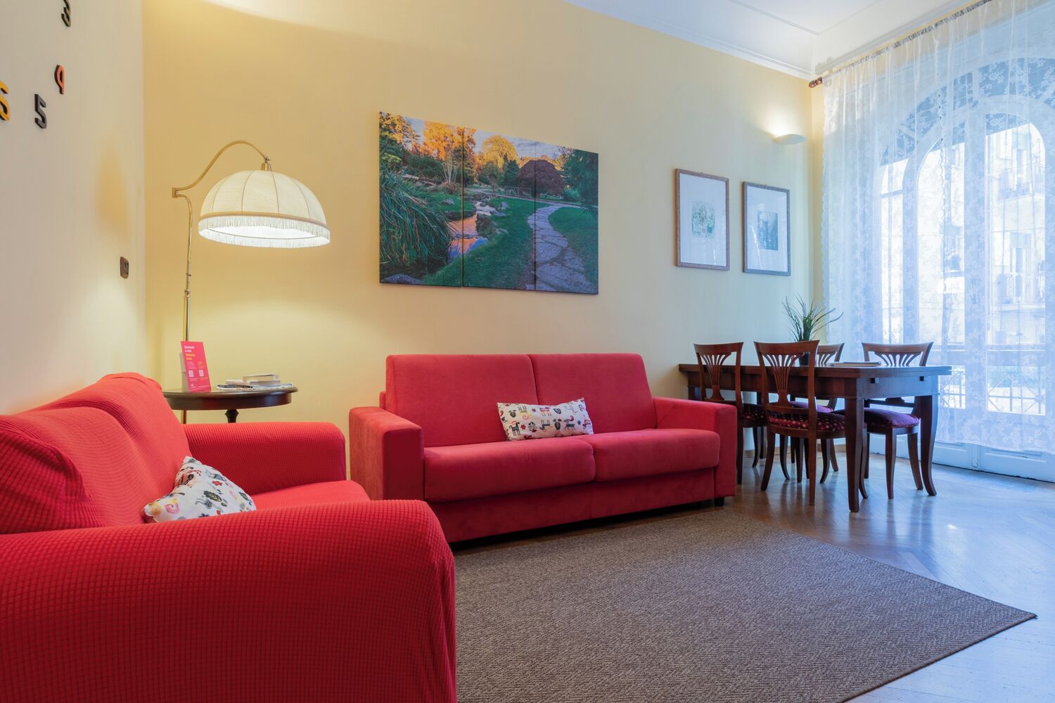 A living room with a red sofa, a lamp, a painting, and a dining area with chairs and a table.