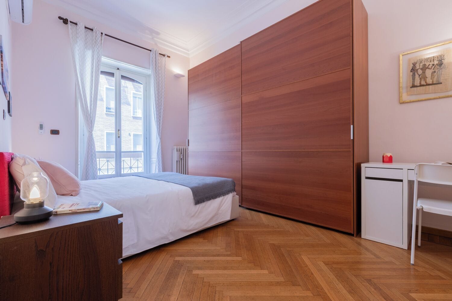 A bedroom with a bed, bedside table, lamp, and a view of the outside through a window.
