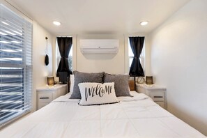 Ein Schlafzimmer mit einem Bett, zwei Nachttischen, einer Wandklimaanlage und einem Fenster mit Jalousien.