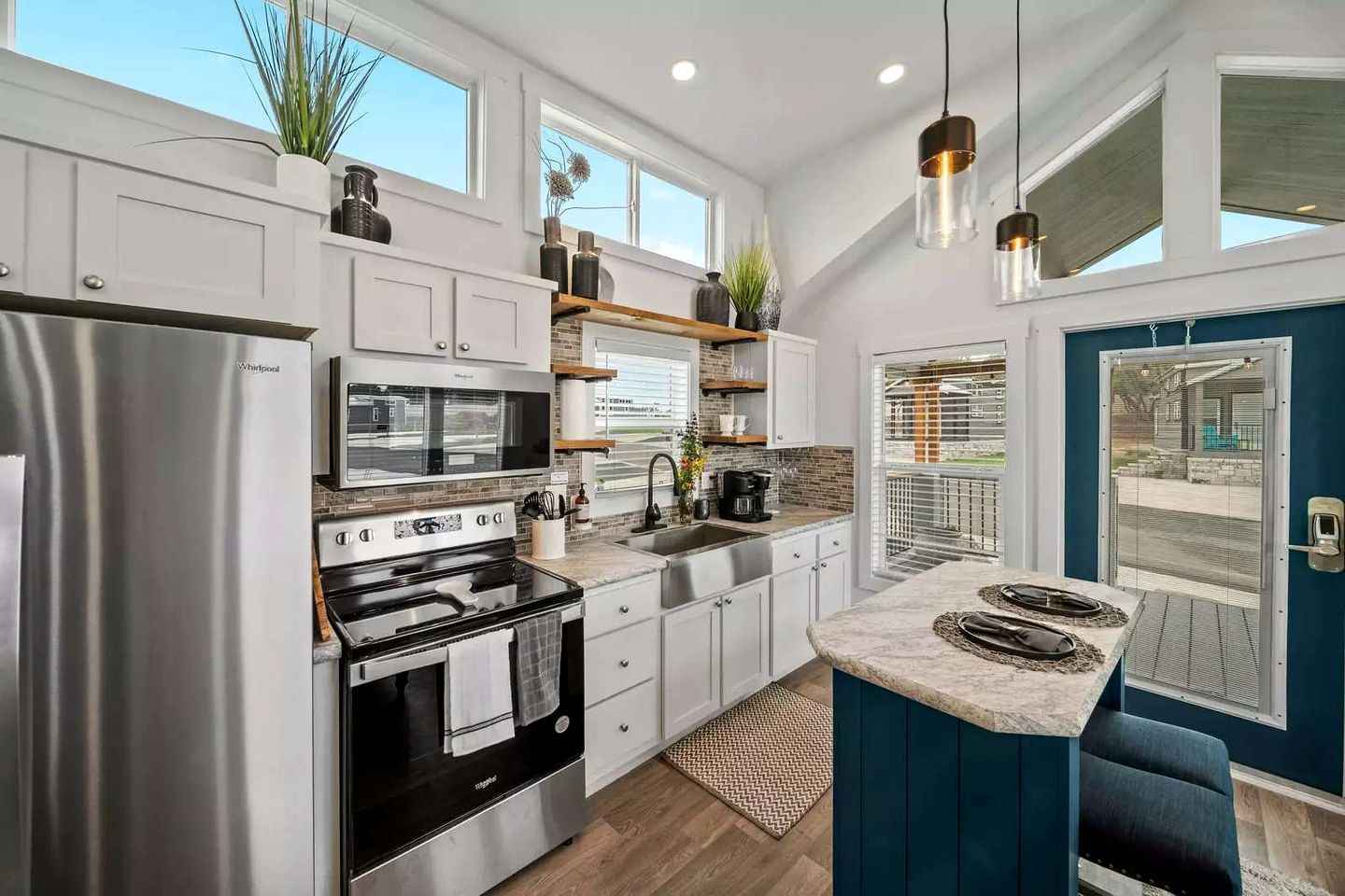 Una cocina moderna con un refrigerador de acero inoxidable, un horno empotrado y un microondas. Hay un lavamanos con grifo y una ventana encima. La cocina tiene armarios blancos y una pared de acento azul.
