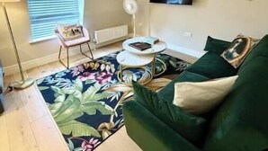 Ein Wohnzimmer mit einem grünen Sofa, einem Couchtisch, einer Stehlampe, einem an der Wand montierten Fernseher und einem Fenster mit Jalousien.