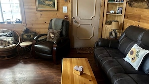A cozy room with a leather sofa, a wooden coffee table, a chair with a cushion, and a small round table with a lamp. - Peaceful Creekside Cabin with Fire Pit & Scenic Deck in Cascade, Idaho (Cascade)