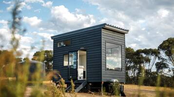 Une cabane moderne aux couleurs sombres, dotée d’un petit porche, d’une grande fenêtre et d’une porte.