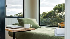 Een slaapkamer met een bed, een raam met uitzicht op bomen, een laptop en een witte mok op een houten tafel.
