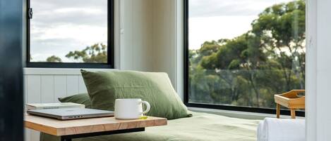 A bedroom with a bed, a window with a view of trees, a laptop, and a white mug on a wooden table.