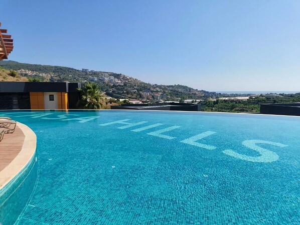 A clear blue infinity pool with 'HILLS' written on the edge, overlooking a hilly landscape.