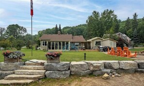 A residential property with a stone retaining wall, a lawn, and a house with a covered patio area. - Waterfront Family Cottage with Hot Tub on Gull River, Ontario (Kawartha Lakes)